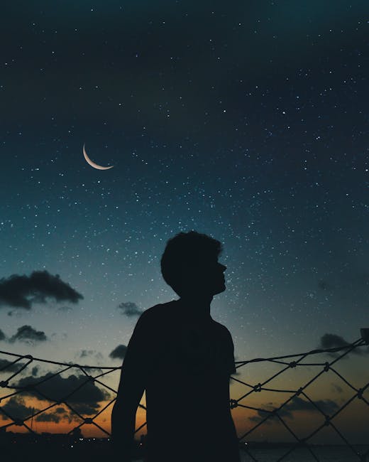 About A lone silhouette stands beneath a crescent moon and starry sky, evoking a sense of solitude and wonder.